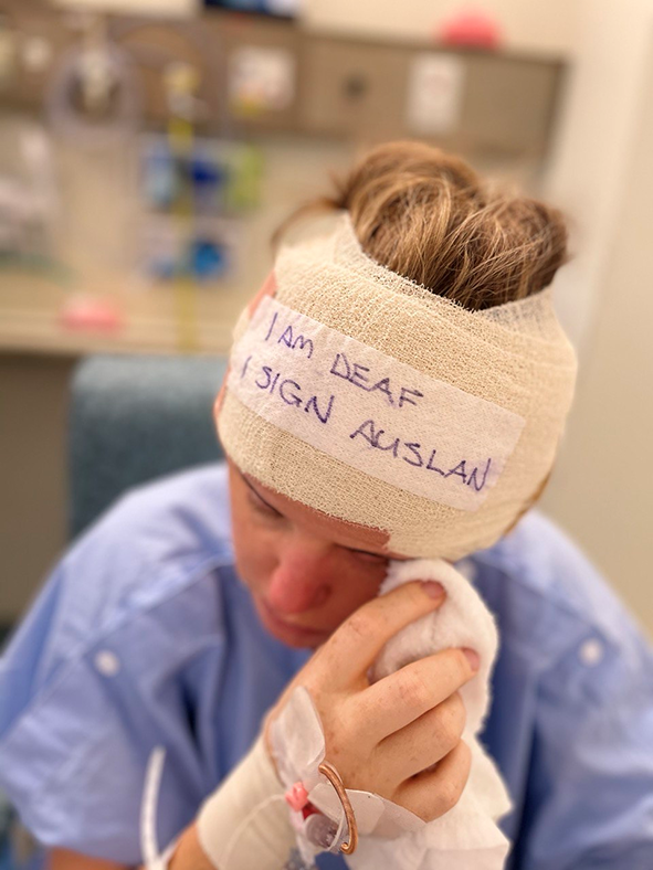 A young woman is wearing a surgical gown after surgery, with a sticker on her head bandage that says "I am deaf, I sign Auslan".